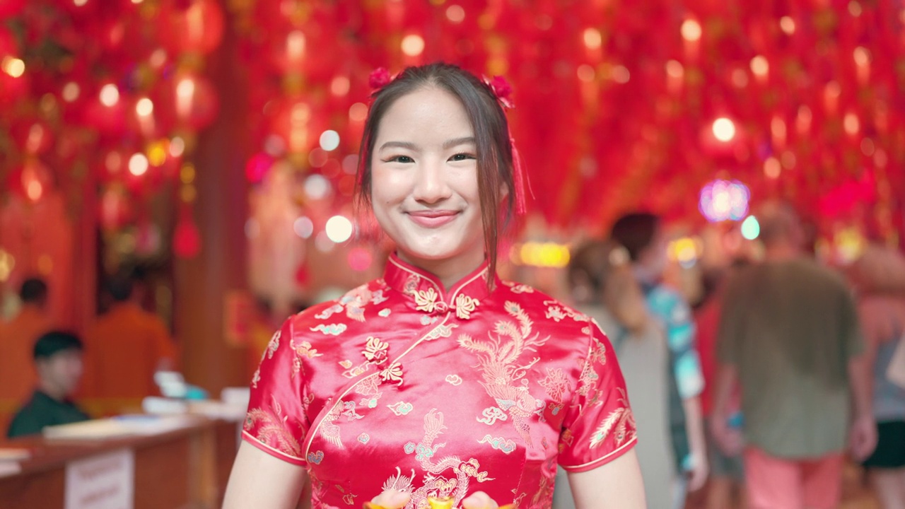 穿着红色旗袍的年轻亚洲女孩给你她手里拿着的金元宝。笑得很开心，看了看镜头。中国新年的概念。视频下载