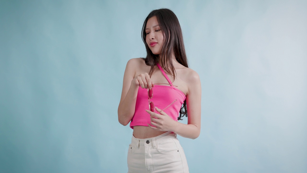 照片中，美丽的亚洲年轻女性微笑着，穿着粉色上衣和白色牛仔裤，站在孤立的蓝色背景上视频下载