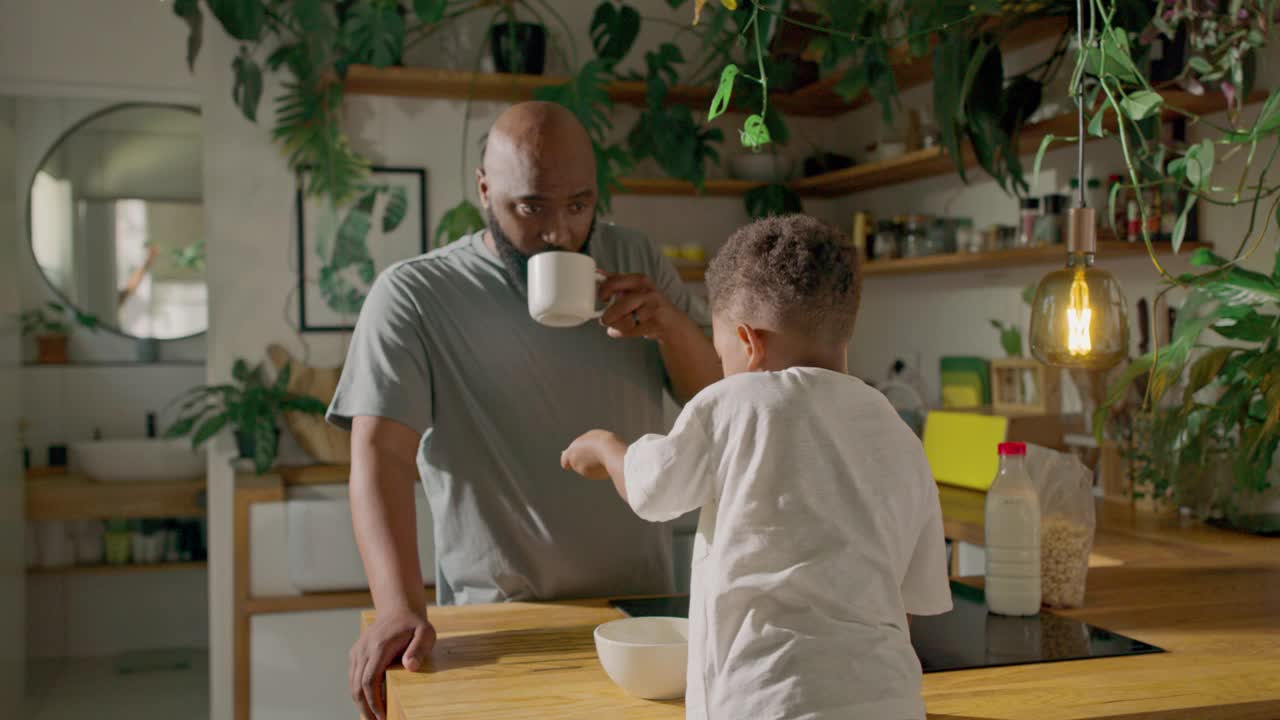 一个充满玩笑和温柔关怀的早晨，一位父亲在绿色装饰的厨房里为儿子准备了特别的早餐时间视频下载