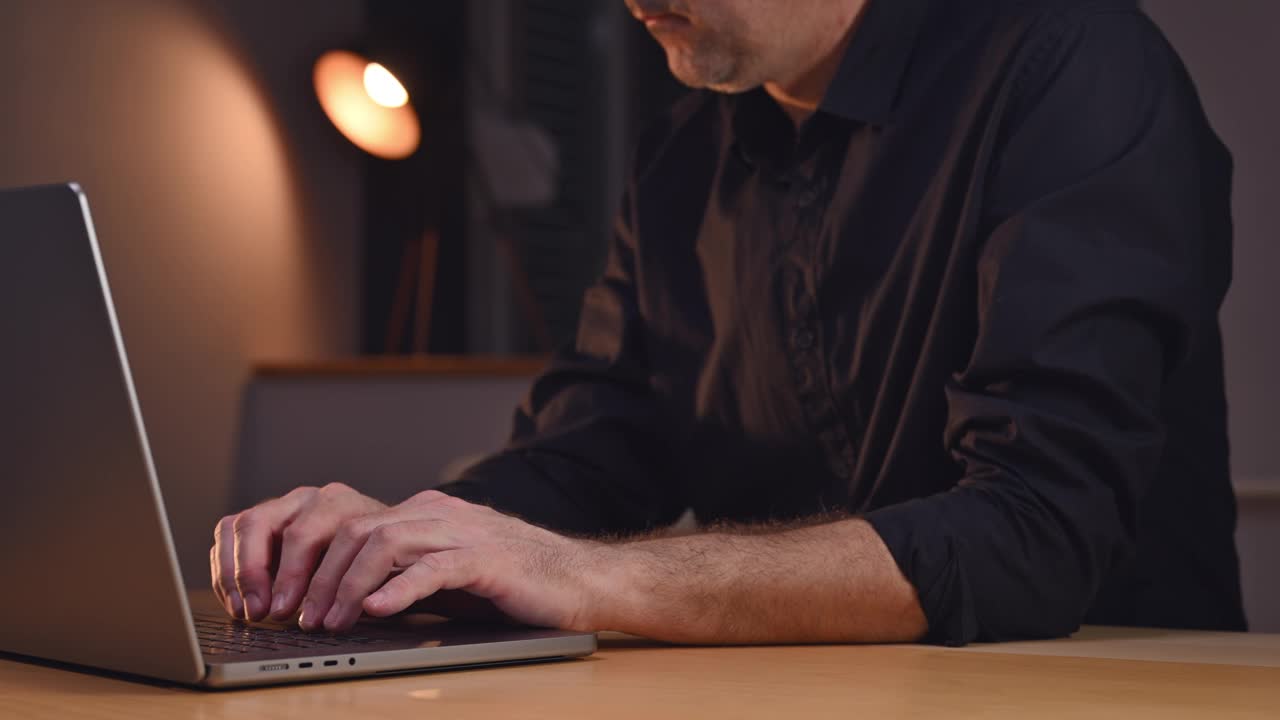 一位穿着休闲服的商人坐在室内，全神贯注地摆弄着个人笔记本电脑，在电子设备键盘上打字视频下载