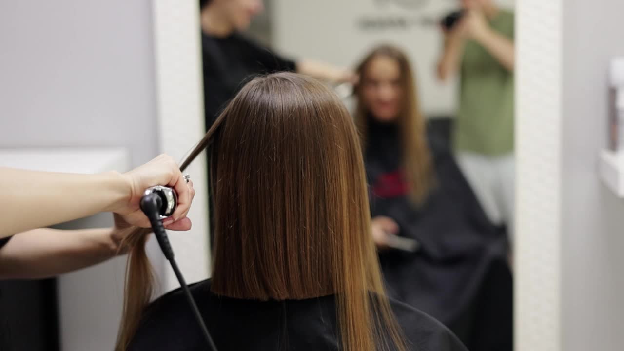 不知名的专业发型师用直发器和梳子为女性客户烫发，在美容院做出时尚的发型。角蛋白，贴膜，头发护理，美发沙龙和发型设计。视频下载