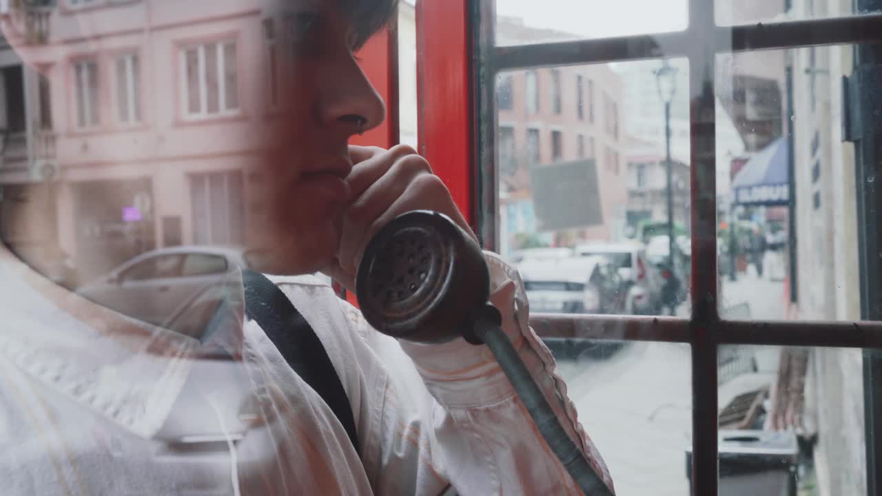 穿着90年代风格的年轻时尚男子在城市街道上的红色复古老式电话亭或盒子里说话视频下载