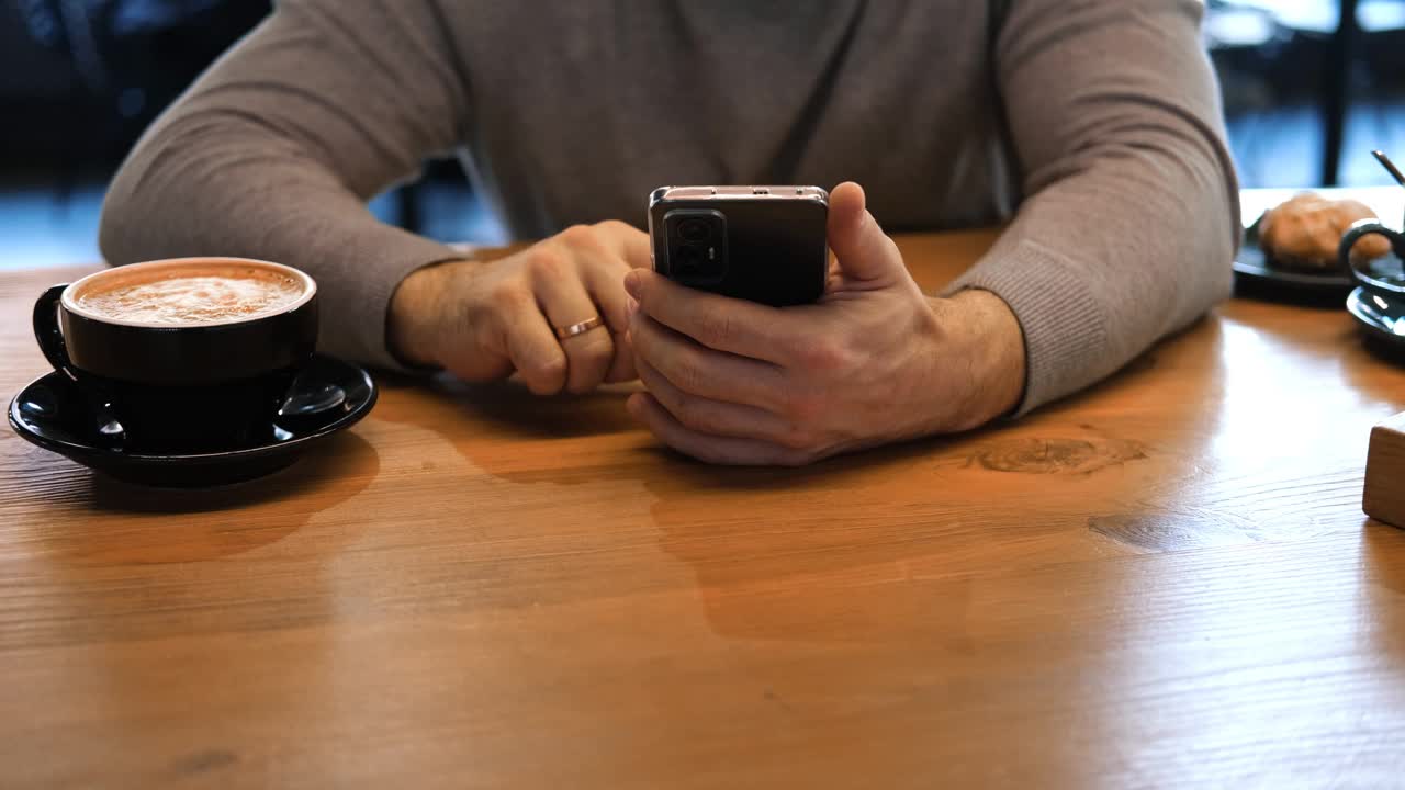 穿着灰色毛衣的成年男子坐在咖啡馆的木桌前玩智能手机。自由职业者的休息时间。时尚男士在舒适的咖啡馆氛围中享受咖啡和电话时光。自由职业、休闲视频下载