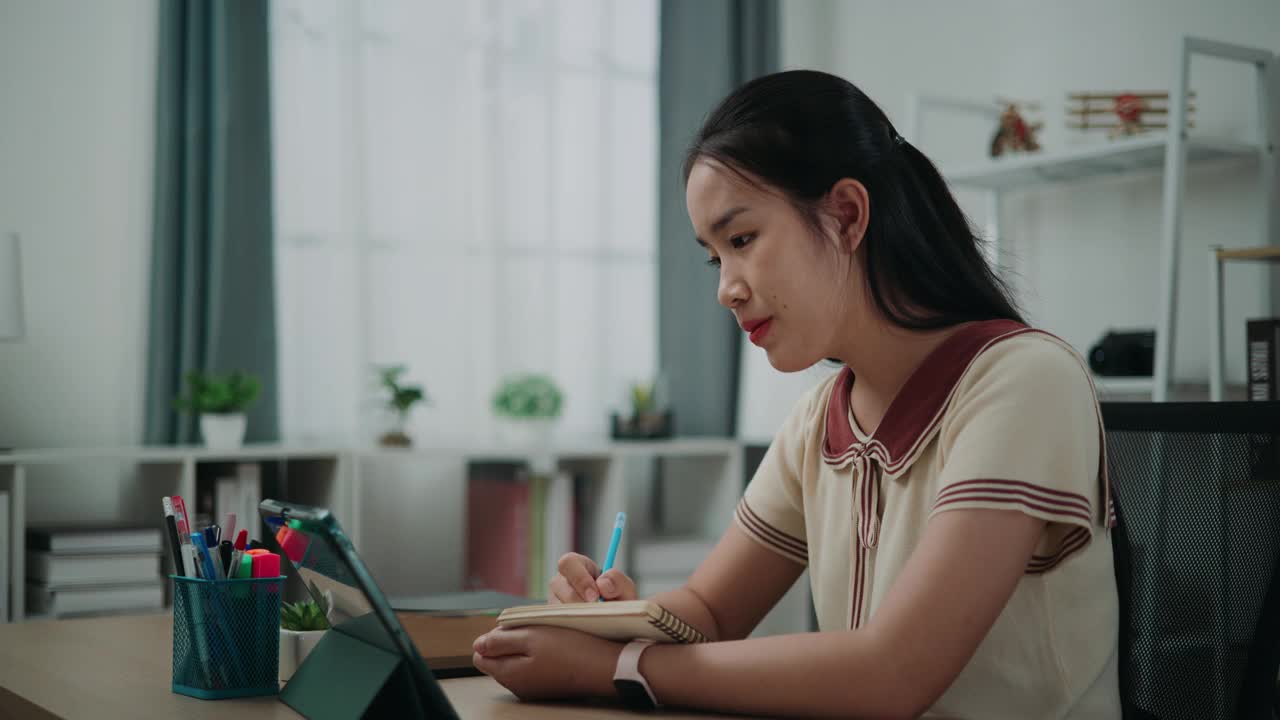 快乐的年轻亚洲女性坐在办公桌上写笔记，同时在家里用平板电脑进行在线学习。视频素材