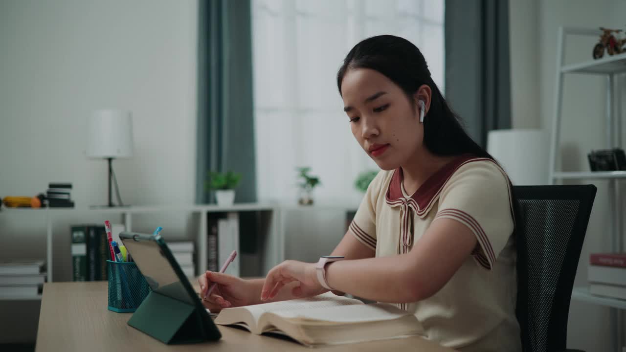 年轻的亚洲女性戴着无线设备坐在书桌前，一边在笔记本上写信息，一边在家用手机看书和工作。视频下载