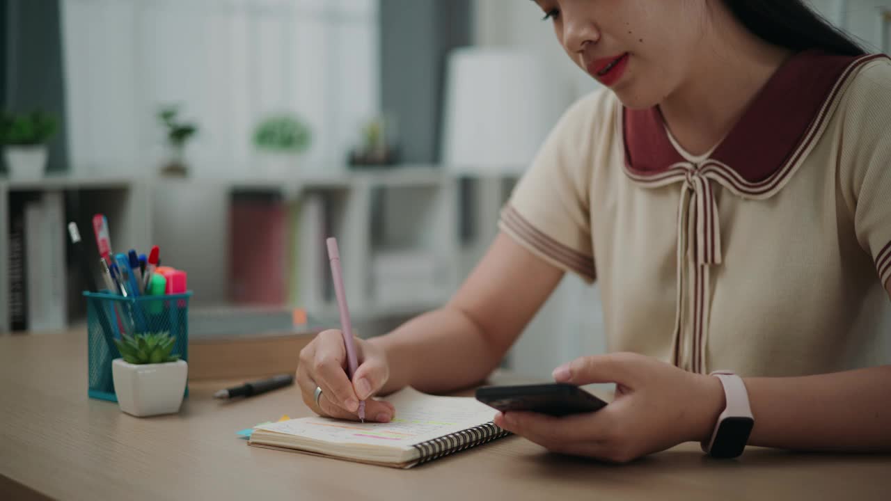 快乐的年轻亚洲女性坐在办公桌前，在家里用智能手机写日记。视频素材