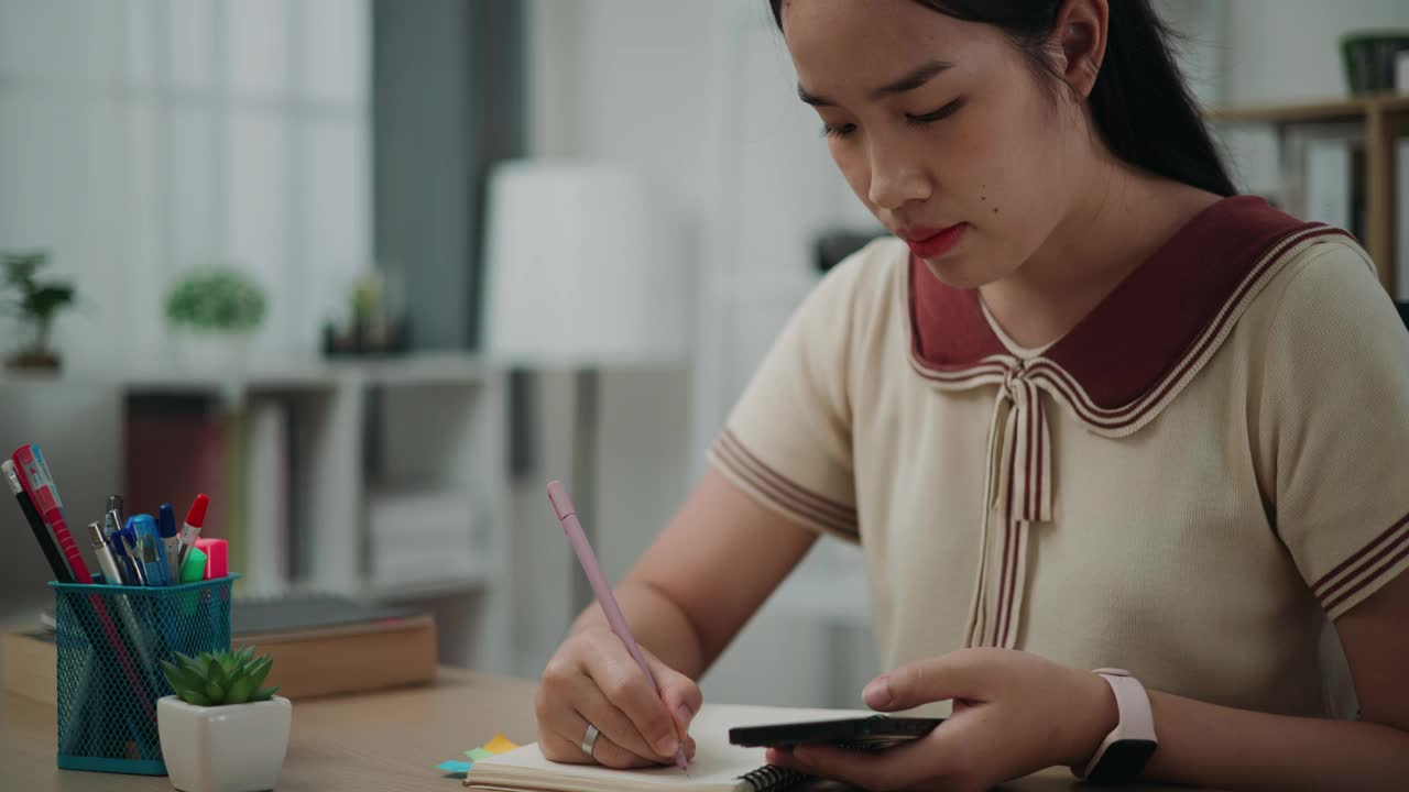 年轻的亚洲女性坐在办公桌前，在家里用智能手机写笔记信息。视频素材