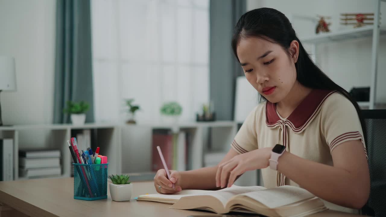 年轻的亚洲女性坐在书桌前，一边在家看书，一边在日记本上写信息。视频素材