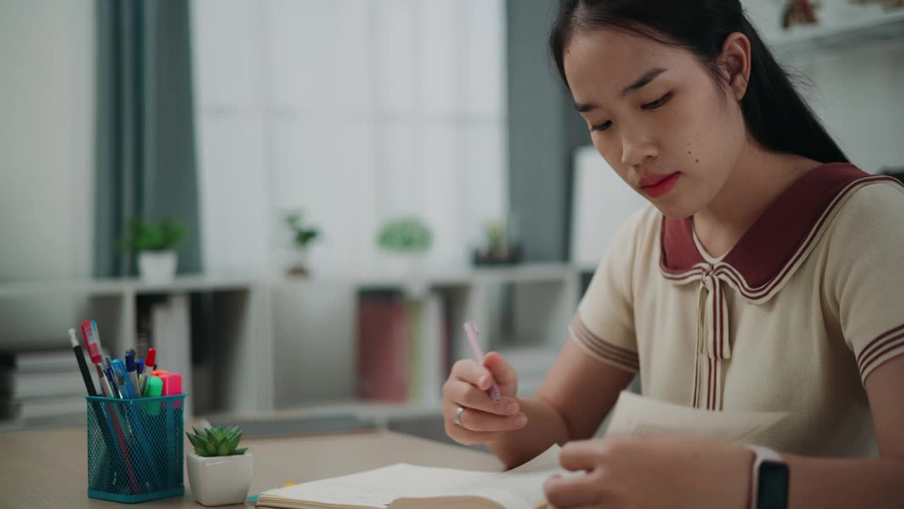 年轻的亚洲女性坐在书桌前，一边在家看书，一边在日记本上写信息。视频素材