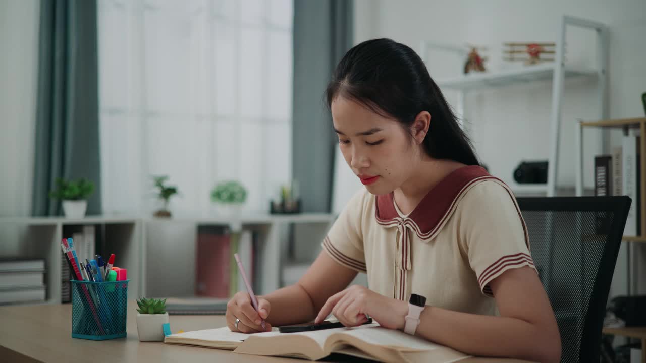 年轻的亚洲女性坐在书桌前，一边在笔记本上写信息，一边在家看书和用手机工作。视频素材