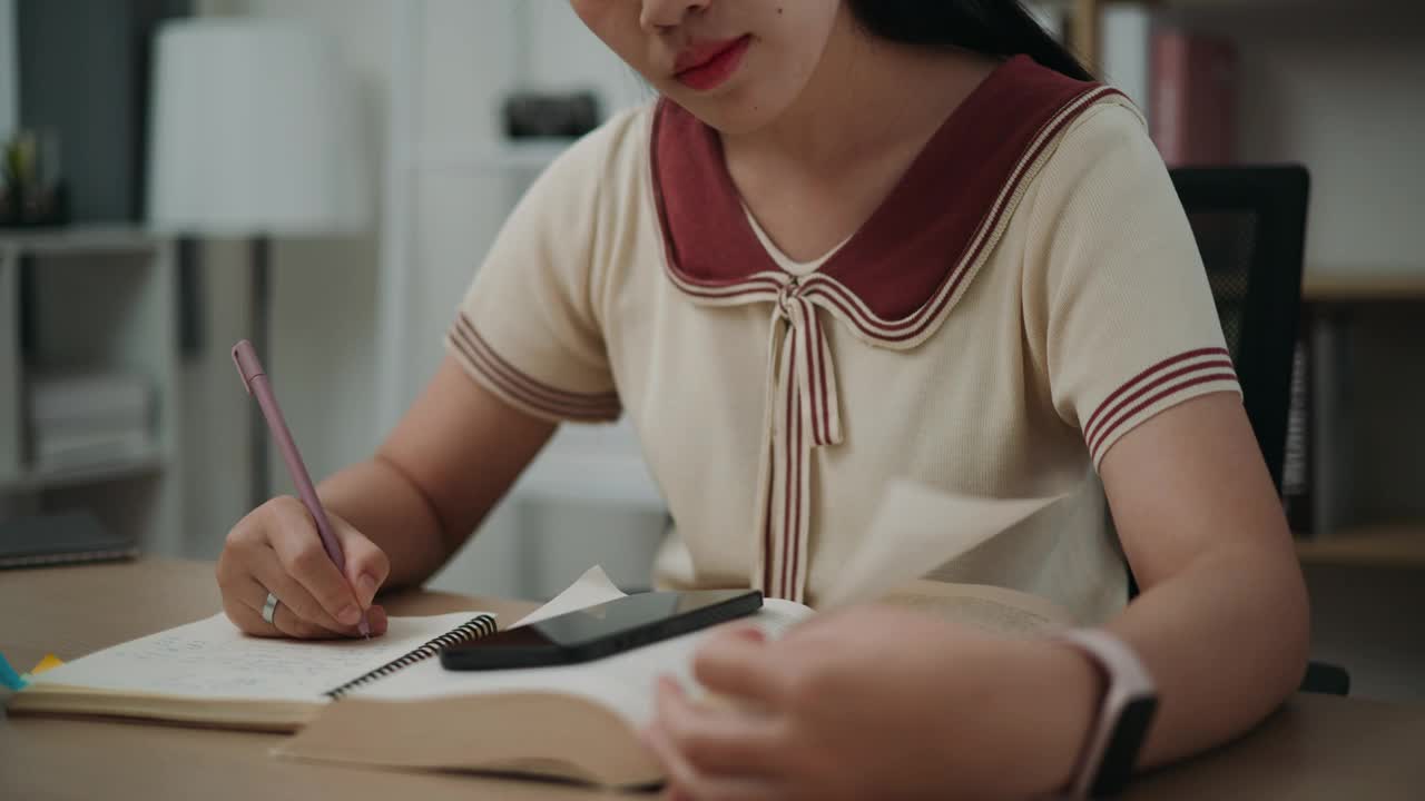 年轻的亚洲女性坐在书桌前，一边在笔记本上写信息，一边在家看书和用手机工作。视频素材
