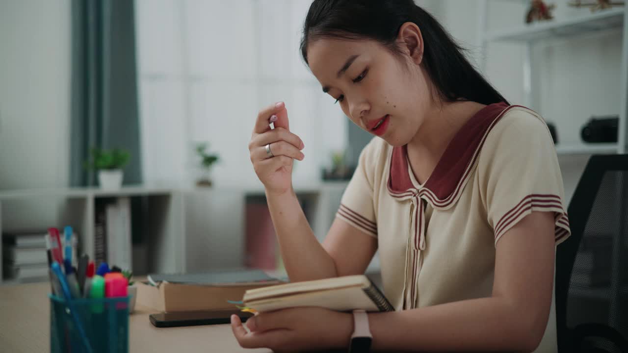 快乐的年轻亚洲女性坐在办公桌前，一边思考，一边拿着笔在家里写日记。视频素材