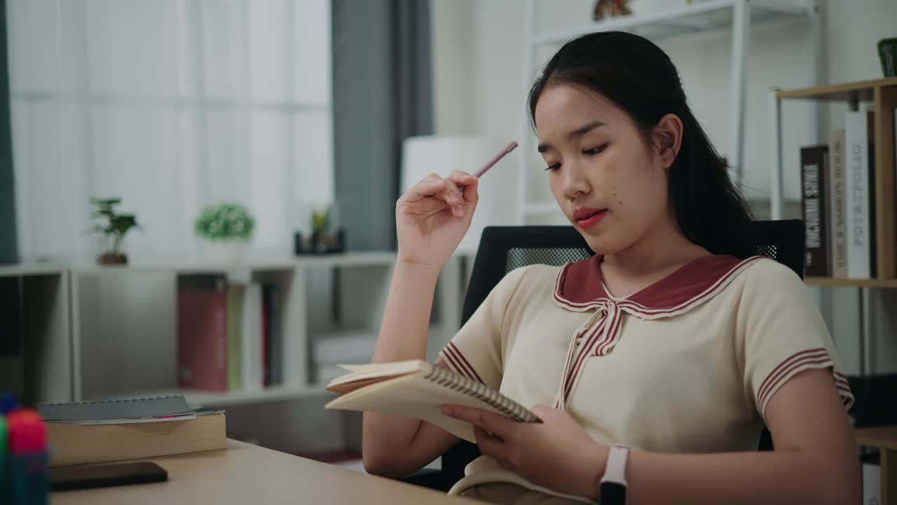 快乐的年轻亚洲女性坐在办公桌前，一边思考，一边拿着笔在家里写日记。视频下载