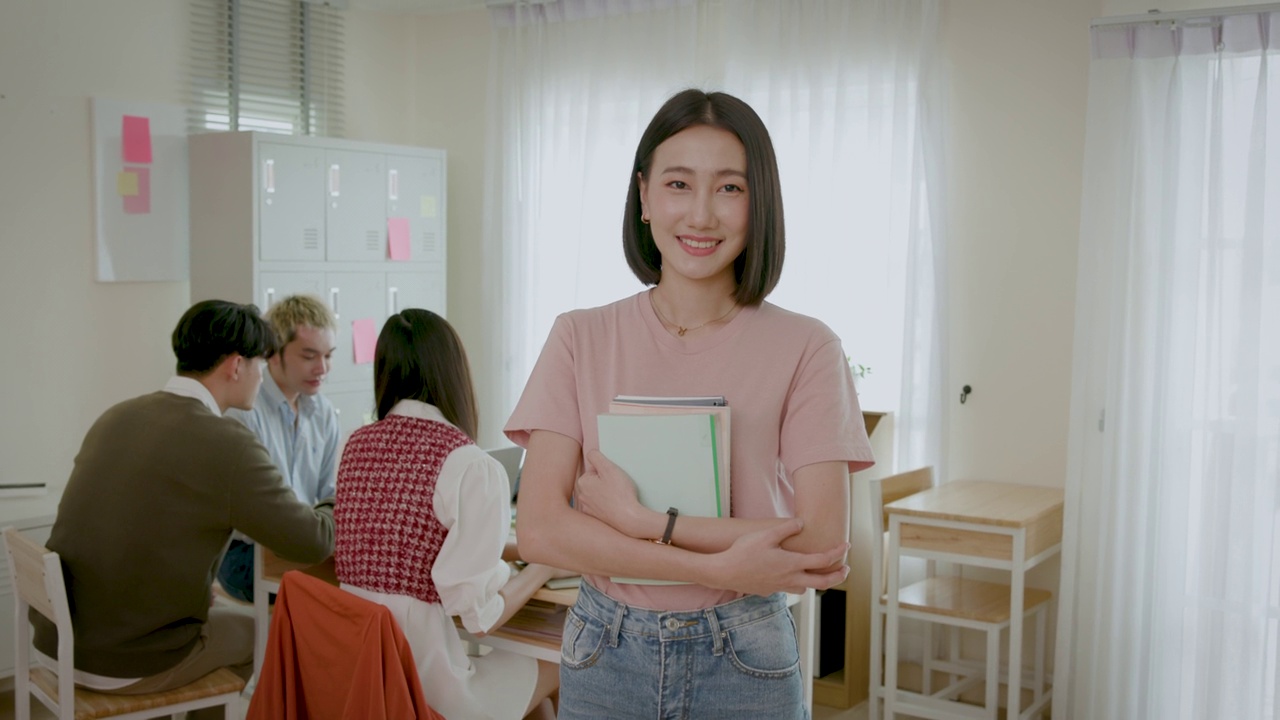 年轻的亚洲人的肖像女学生拿着书站在教室里，而她的朋友在后台学习视频下载