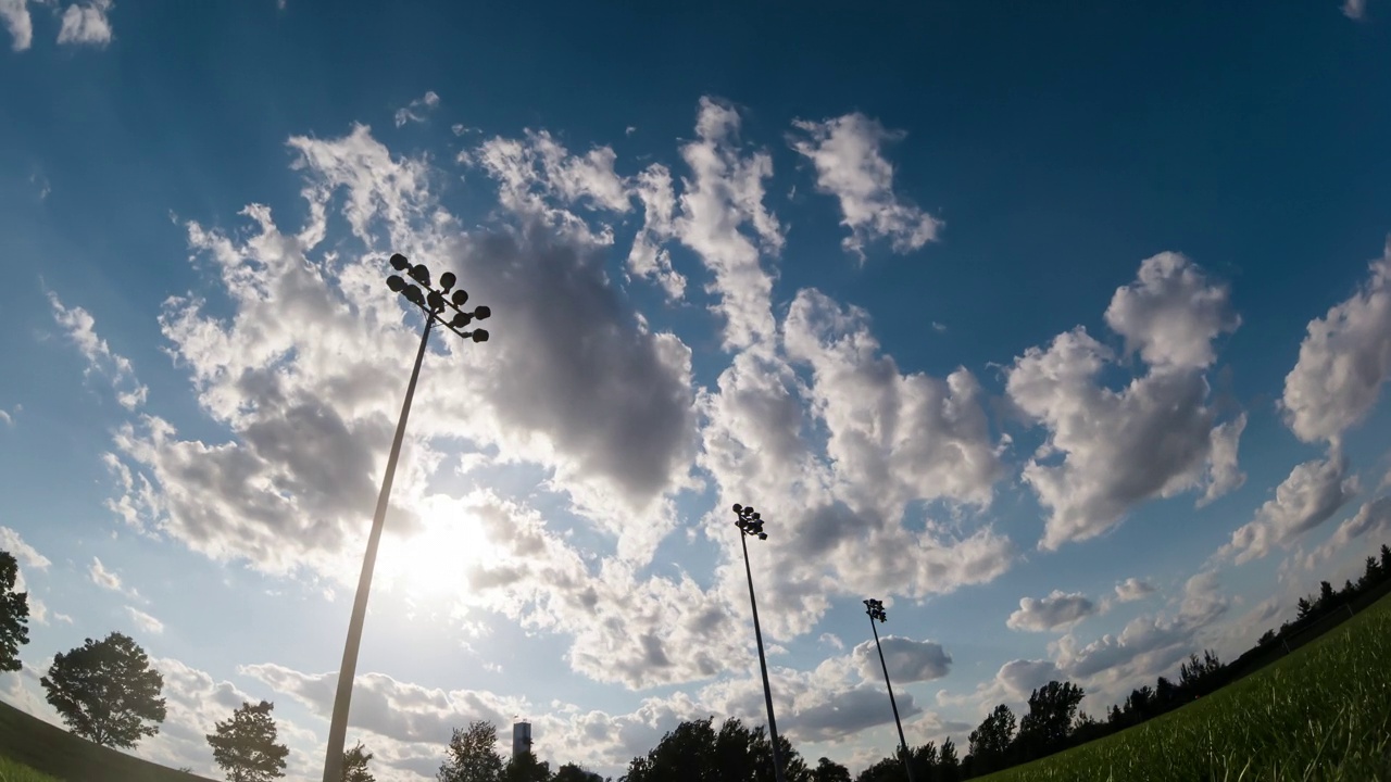 随着时间的流逝，夕阳和夏日的云彩映衬着足球场的灯塔，天空快速而流畅的移动。足球场的手电筒和晚上的时间流逝。视频下载