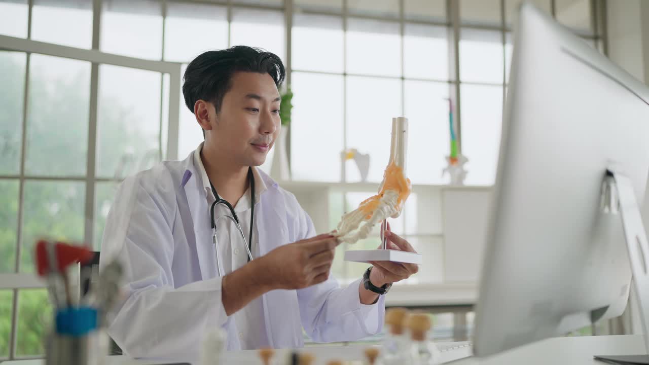 在线医疗咨询。亚洲男骨科医生展示足部骨骼、踝关节解剖模型，通过在线通话、视频通话为患者讲解。理疗，医疗视频下载