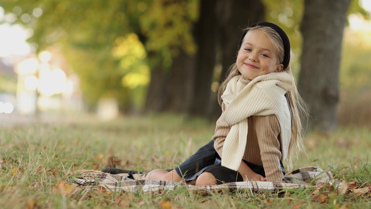 时髦迷人的小女孩高加索5-6岁在户外玩耍，吹落叶从她的手，坐在草地上的格子在秋天的公园户外。儿童节。假期休闲活动视频下载