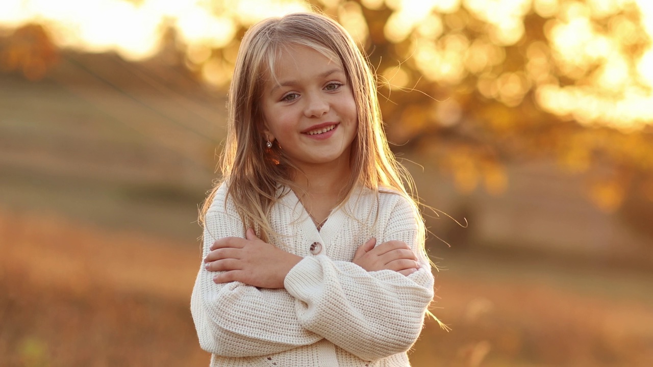 儿童节。可爱的高加索女孩5-6岁的肖像，在秋天的夕阳下，她双手交叉，微笑着看着相机。秋季假期的休闲活动。视频下载
