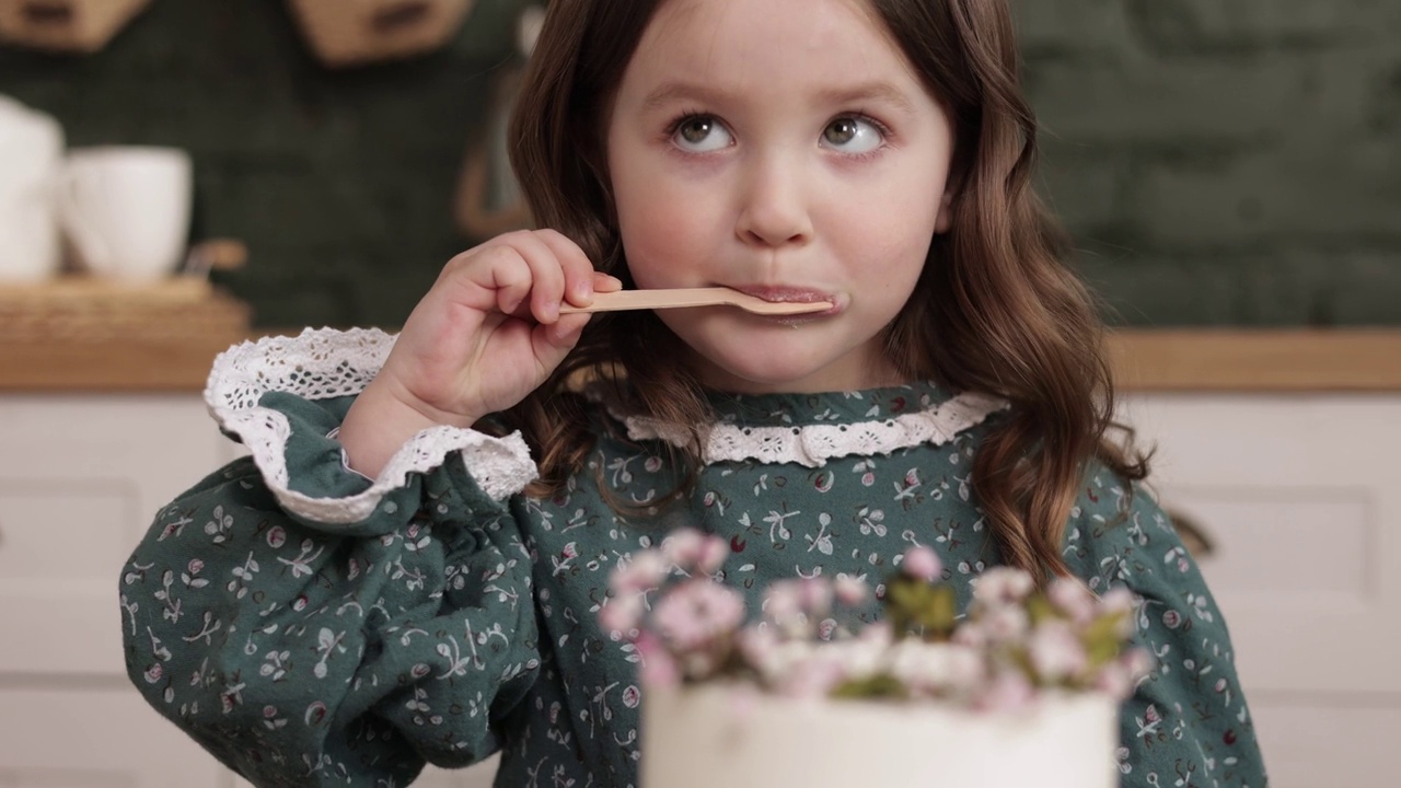 孩子们在家里过生日。可爱的4-5岁的小女孩穿着节日服装，正在开心地吃着装饰着鲜花的生日蛋糕。快乐的孩子用白奶油舔木勺视频下载