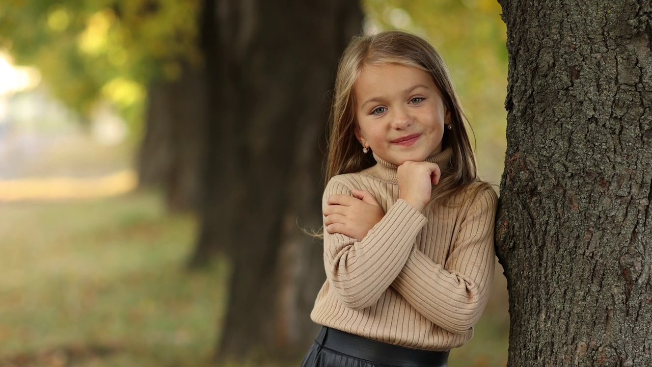 儿童节。5-6岁的开朗小女孩站在秋天公园的树旁，对着相机摆姿势微笑。时尚温柔的学龄前孩子穿着米色针织毛衣。快乐的童年。视频下载