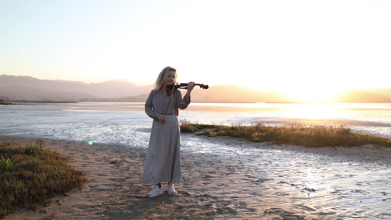 日落时分，金发碧眼的艺术家在海滩上拉小提琴视频下载