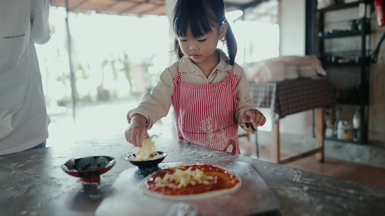 一个小女孩戴着可爱的水桶帽，系着围裙，在烹饪课上用擀面杖把面团擀平，在手工制作披萨时给奶酪浇头。视频下载