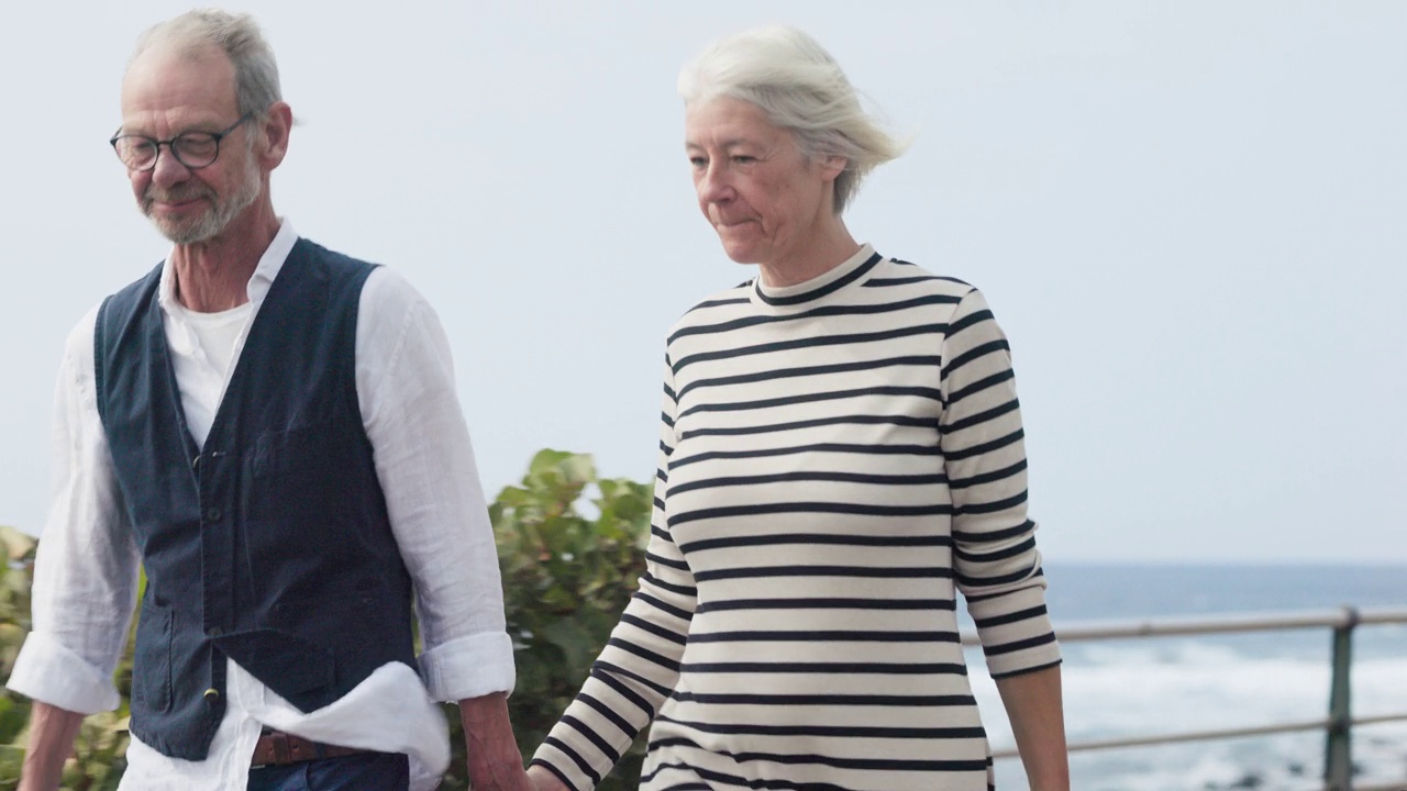 一对老年夫妇正手拉手在海边散步，边走边接吻。视频下载