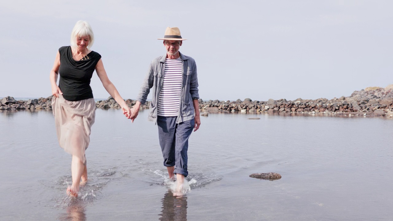一对老年夫妇在阳光明媚的夏日里，手挽着手在海边的泻湖里散步。视频下载
