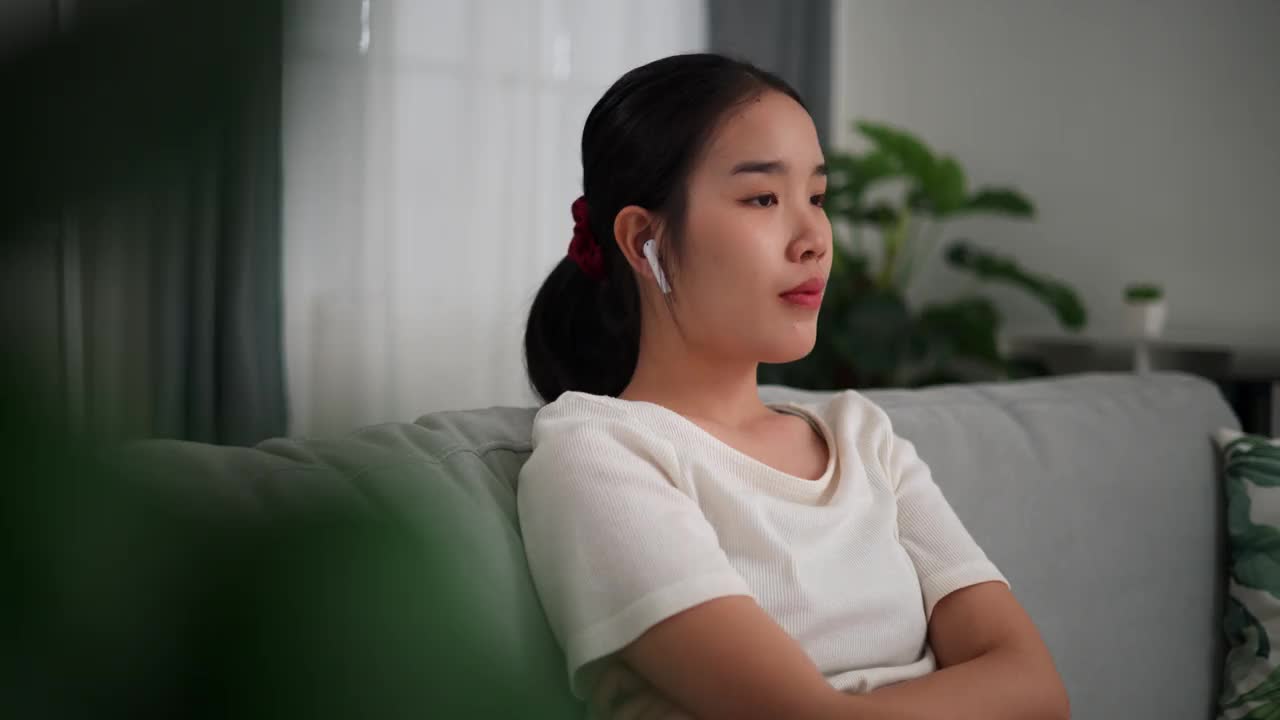 一位年轻女子坐在家里客厅的沙发上，戴着无线耳机，开心地听着音乐。视频下载