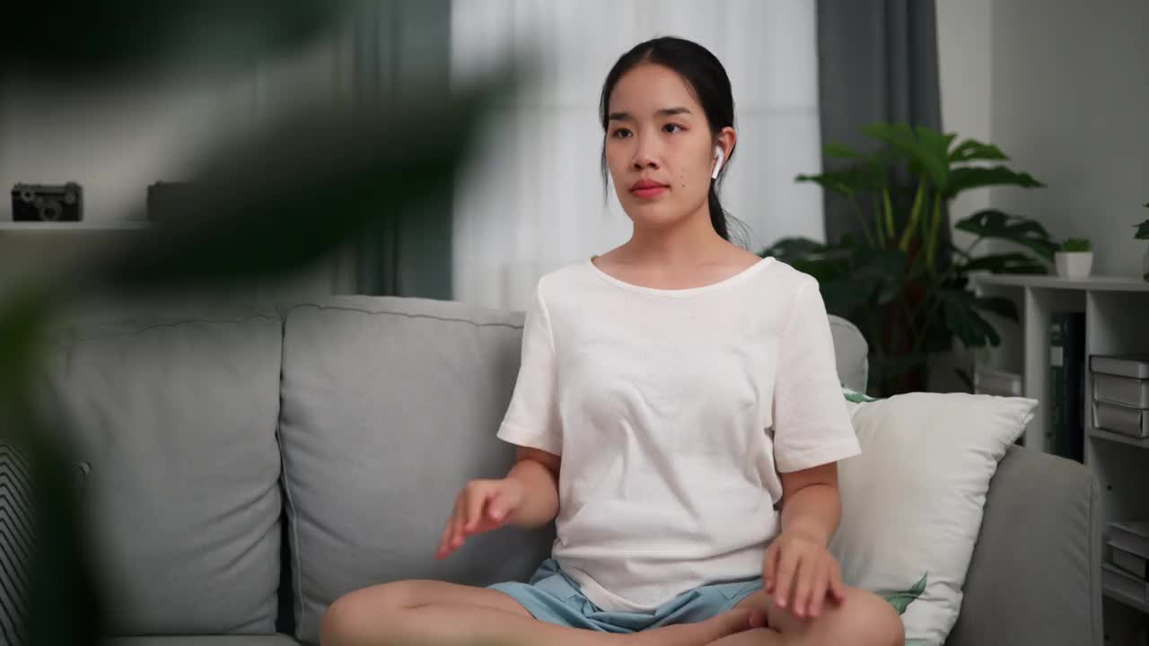 年轻女子在家里客厅的沙发上戴着无线耳机做瑜伽。视频下载
