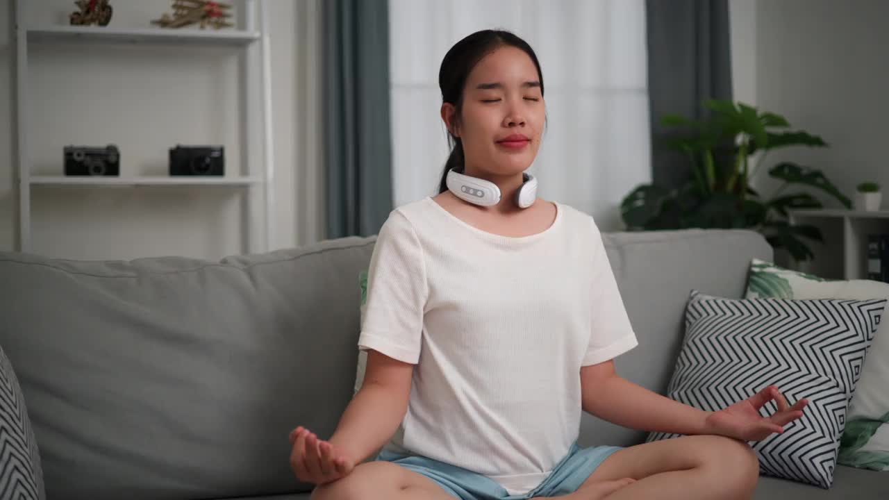 年轻女子坐在家里的沙发上使用颈部按摩器。视频下载