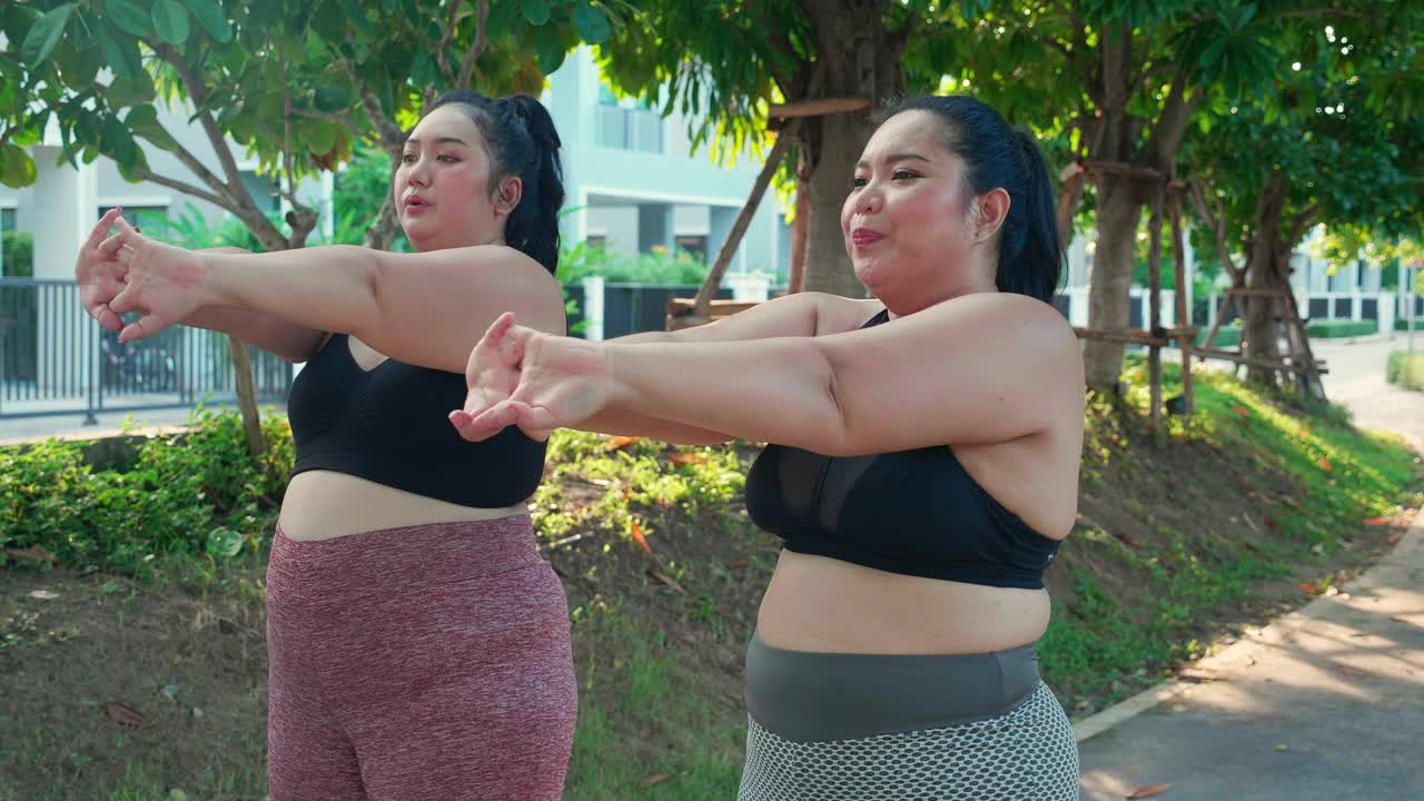 肥胖的两个年轻的亚洲妇女在公园里伸展手臂肌肉，超重的妇女在握手时为节食和健康做热身和锻炼，超大的和锻炼，加上尺寸。视频下载