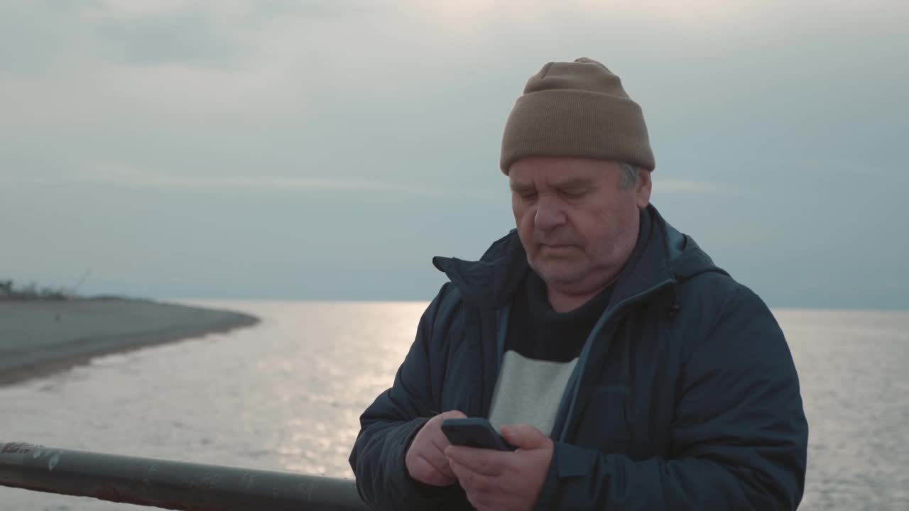 一个成熟的男人站在码头上全神贯注地玩手机，黄昏的大海背景是平静的视频下载