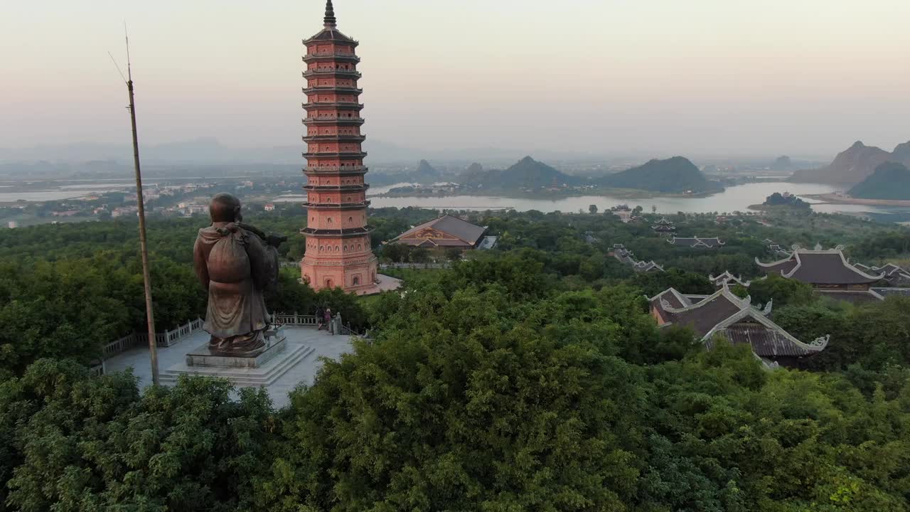 日落时，在越南宁平蛇河前的佛寺、宝塔和长满绿树的佛像上空的无人机鸟瞰图视频素材