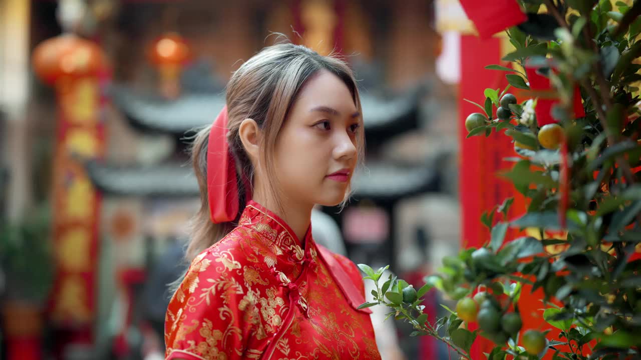 年轻的亚洲女孩在中国寺庙里穿着红色旗袍微笑的画像，寺庙里装饰着纸红灯笼。视频下载
