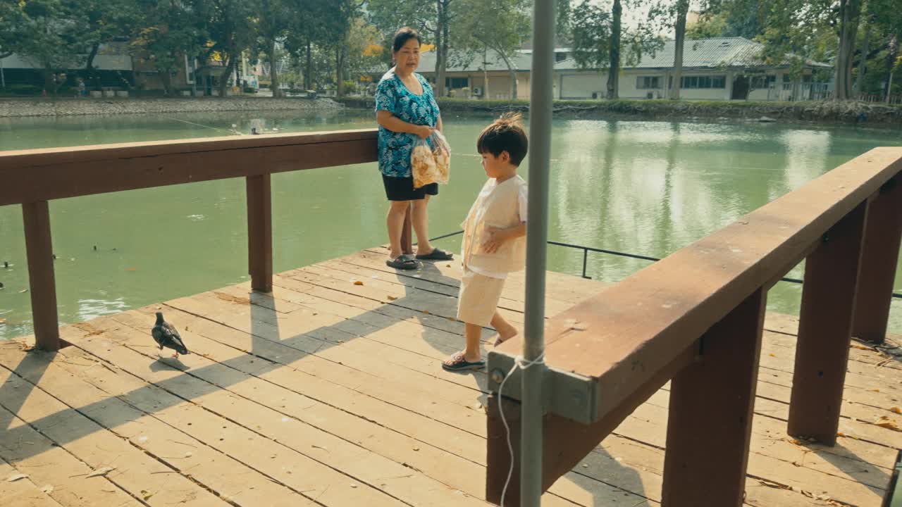 亚洲奶奶和孙子在宁静的池塘里喂鱼，在大自然的美丽中捕捉爱和联系。视频下载