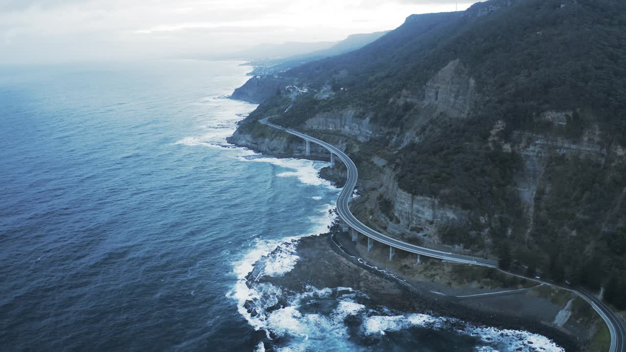 从空中俯瞰汽车在海崖桥上行驶，无人机在波涛汹涌的海面上向后飞行——澳大利亚卧龙岗视频素材
