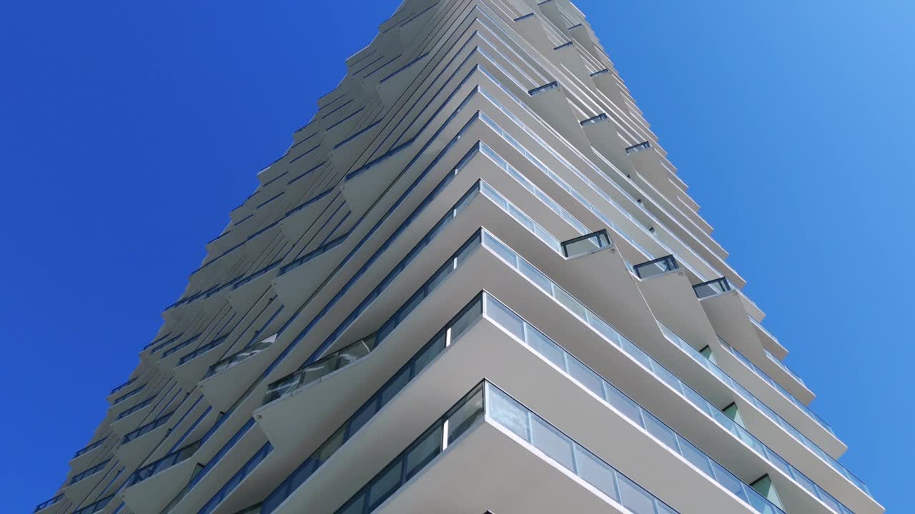 现代摩天大楼的底部视图，具有吸引人的建筑设计，背景是清澈的蓝天。有多个阳台的现代化高层酒店视频下载