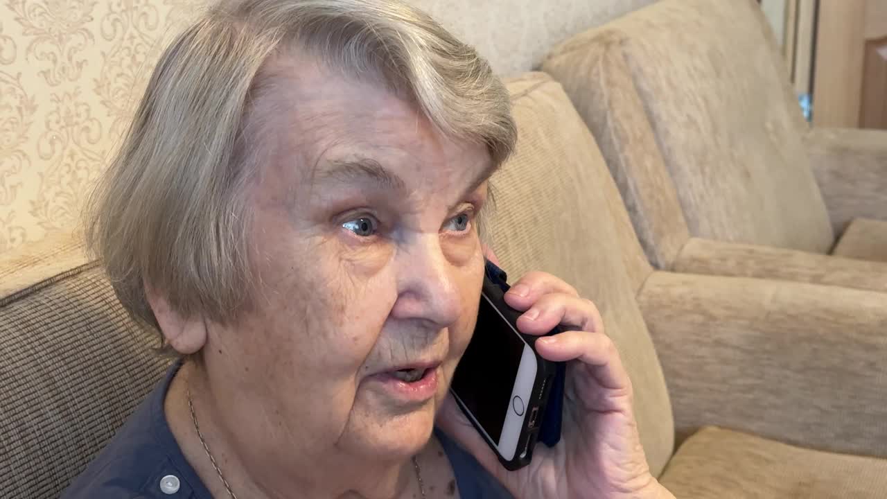 一位80多岁的老太太坐在米色沙发上，手里拿着智能手机，在家里用智能手机和家人愉快地聊天。退休的成年祖母在用智能手机聊天视频下载