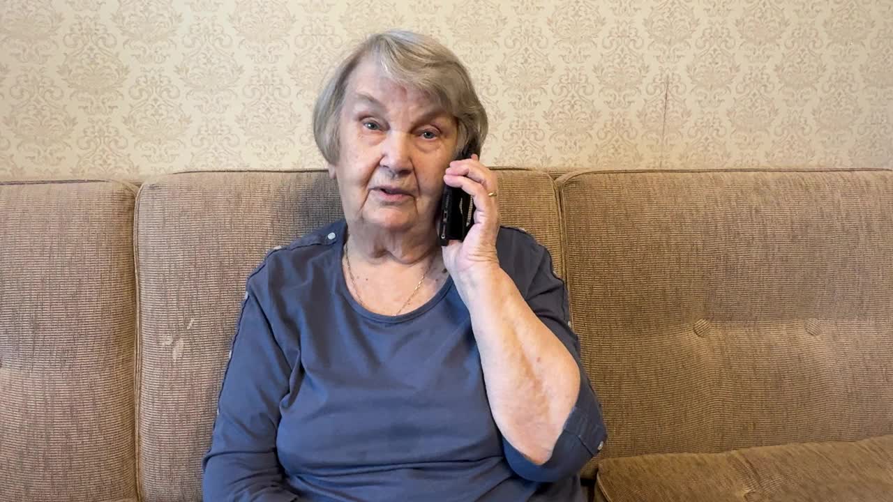 一位退休的成年祖母看着镜头，用她的智能手机说话。一位80多岁的老太太坐在米色沙发上，手里拿着智能手机，开心地和家人聊天。视频下载