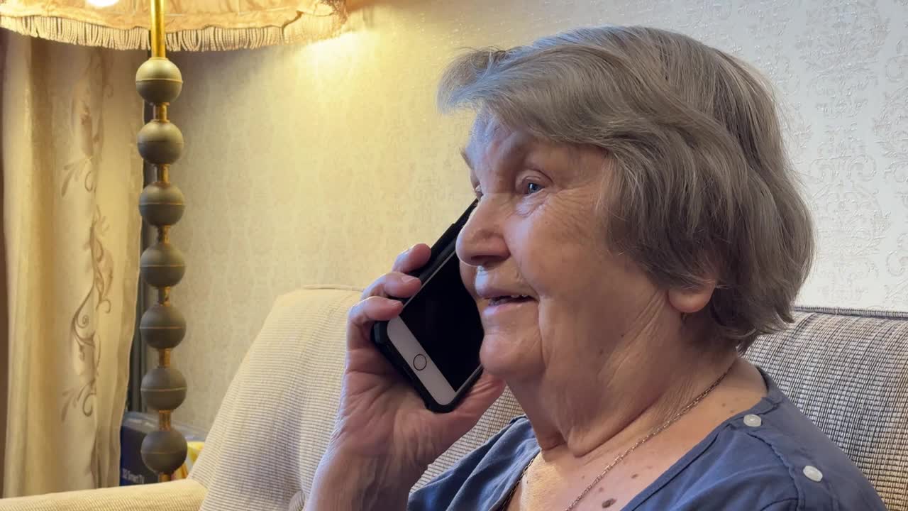 一位80多岁的老太太坐在米色沙发上，手里拿着智能手机，在家里用智能手机和家人愉快地聊天。退休的成年祖母在用智能手机聊天视频下载
