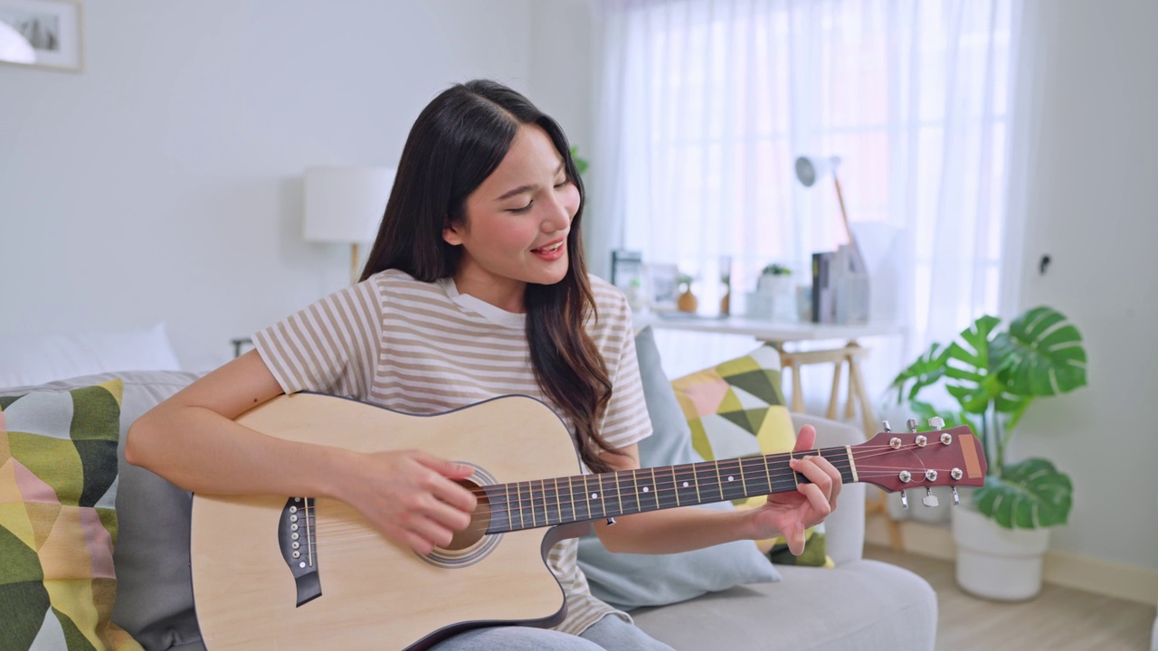 亚洲美女独自在客厅弹吉他。迷人的年轻女孩音乐家感到快乐和放松，坐在沙发上，周末在家练习表演音乐剧。视频下载