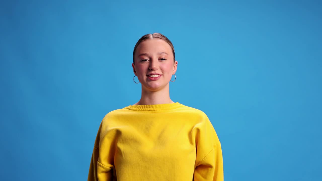 快乐和兴奋的年轻女孩穿着黄色运动衫，在蓝色的工作室背景下微笑着摆姿势。惊喜。视频中,4 k视频下载