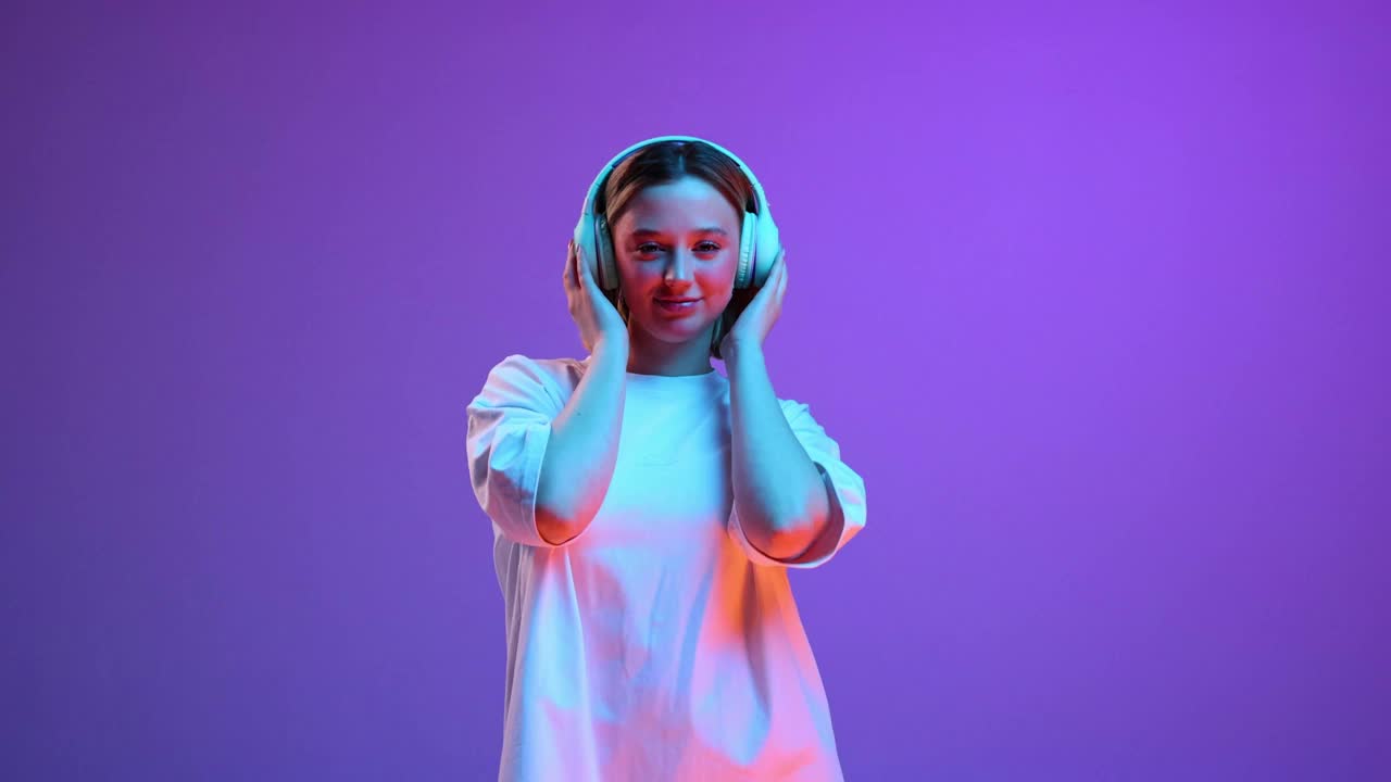 年轻积极的女人穿着休闲白t恤，戴着耳机听着音乐，背景是紫色的霓虹灯。视频中,4 k视频下载