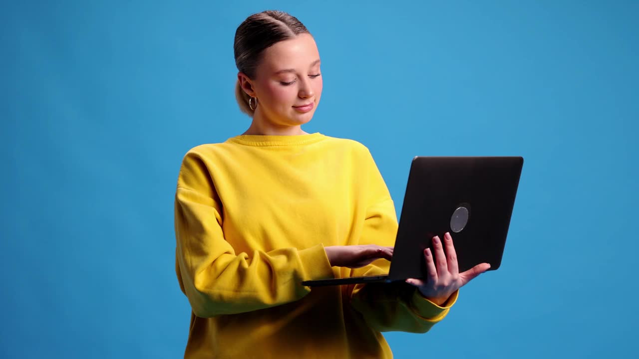 年轻的女孩，穿着黄色运动衫的学生拿着笔记本电脑工作，学习，在蓝色的工作室背景下网购。视频中,4 k视频下载