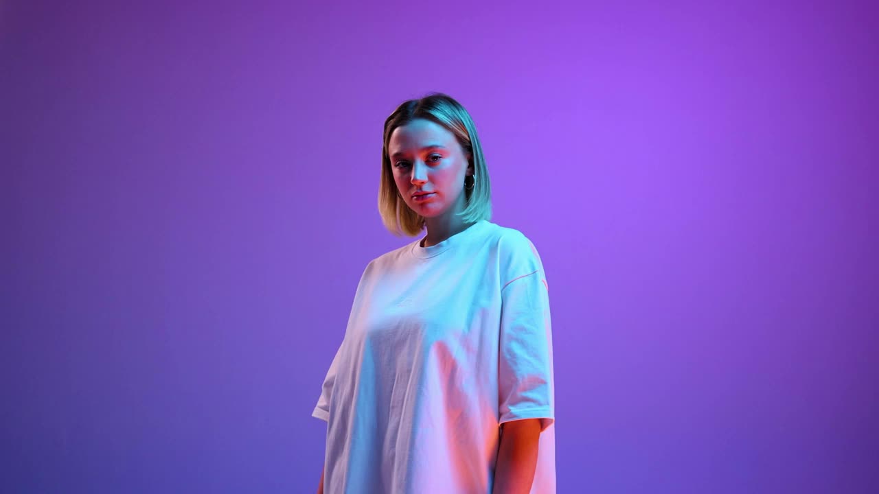 穿着白色t恤的漂亮年轻女孩站着，在霓虹灯的紫色工作室背景下摆姿势。视频中,4 k视频下载