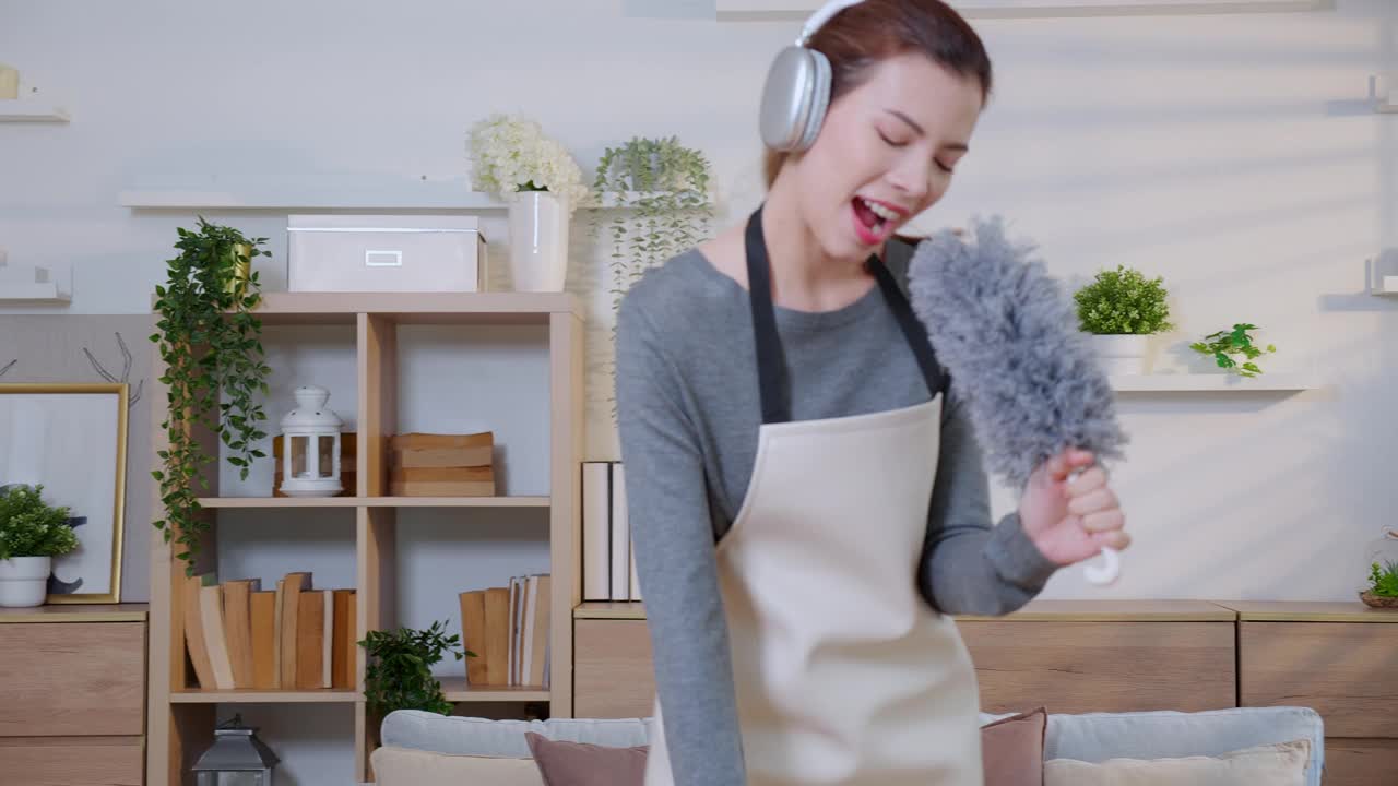 年轻的亚洲女性在明亮整洁的客厅里一边打扫一边戴着耳机听音乐，女佣在家里打扫一边听音乐和唱歌，生活方式概念。视频下载