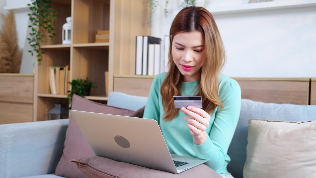 年轻的亚洲女性坐在沙发上用笔记本电脑上网购物，用信用卡购买互联网，女性在沙发上支付，购买和支付，商业和生活方式。视频下载