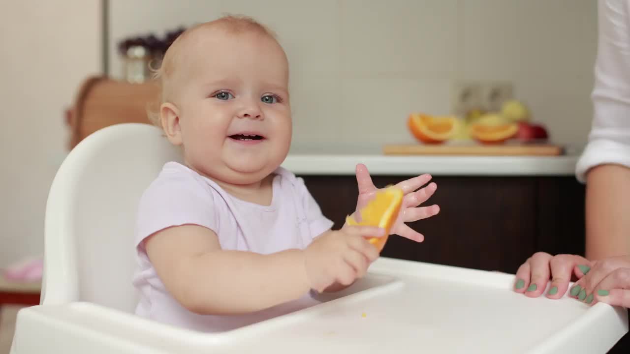 可爱的小女孩在吃橘子。小女儿坐在喂食椅上，吃着厨房里的橘子片，和妈妈一起玩。多汁的水果橙子是健康的饮食。快乐的童年。视频下载