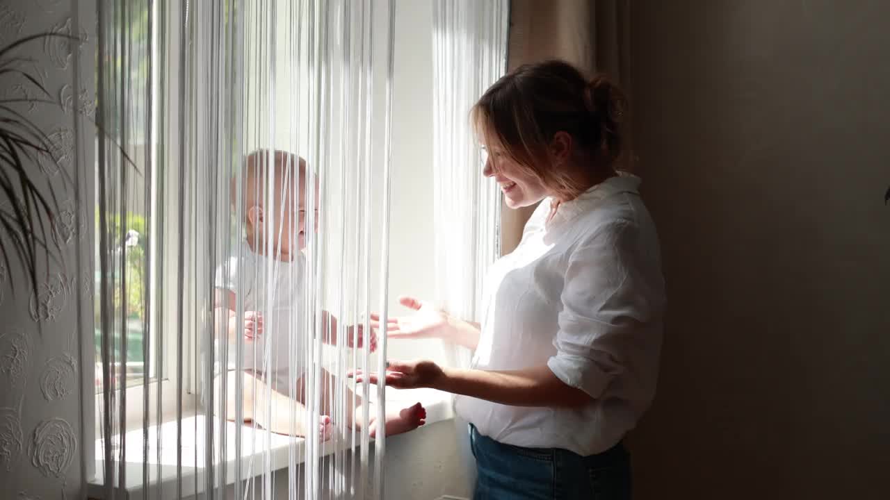 婴儿的女儿或儿子坐在窗台上和妈妈一起玩。快乐的年轻母亲带着女婴或男婴在家里的大窗户旁休息。家庭。孕妇的概念。母亲视频下载